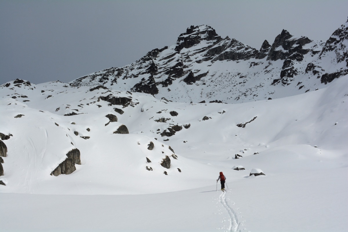 雪山のなだらかな雪原を、バックパックを背負った登山者が奥の岩峰に向かって歩いている風景